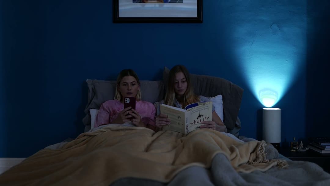 Two women in bed, one on her phone and one reading a book