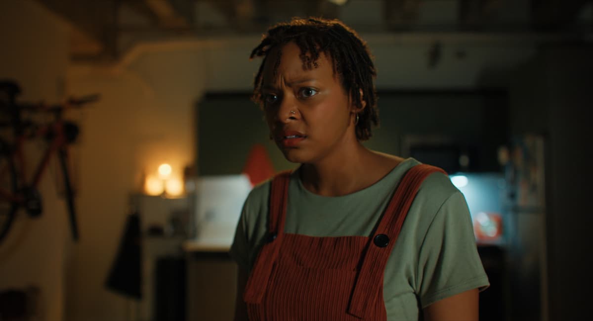 A woman stands looking at something offscreen, a concerned expression on her face.