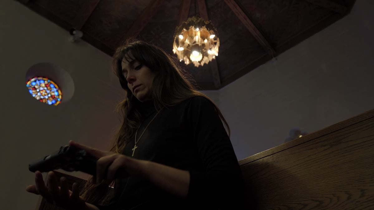 A woman sits in a church, looking down at the pistol in her hand.